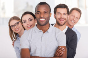 Cheerful young African man keeping arms crossed and smiling while group of people standing behind him in a row and looking at camera